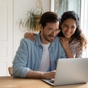 Happy couple use modern laptop at home together Happy Caucasian young couple look at laptop screen browse surf wireless internet on gadget. Smiling millennial man and woman shopping paying on internet on computer at home. Technology concept.